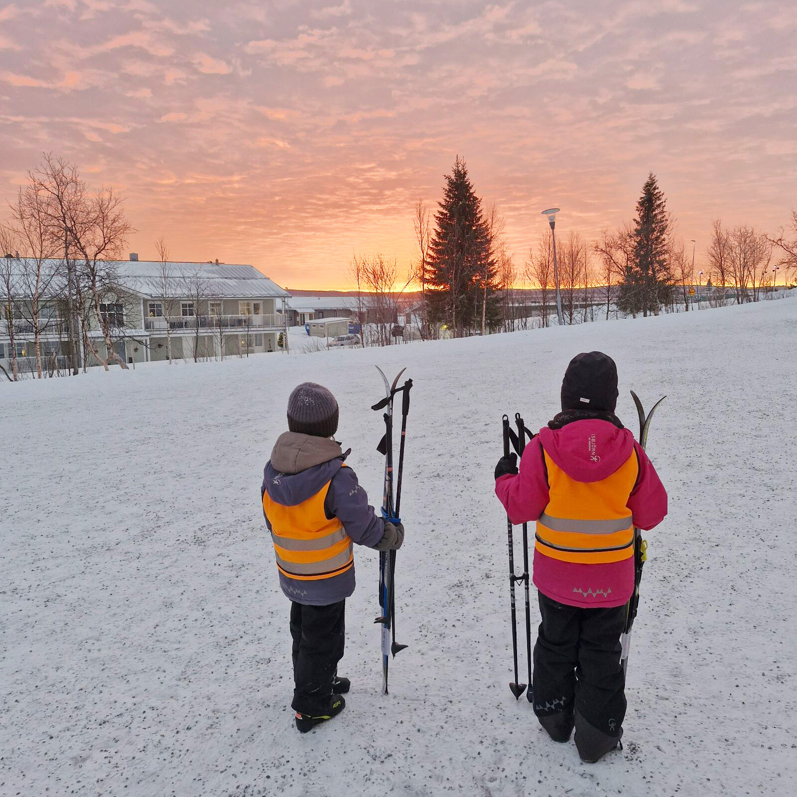 Två barn i reflexvästar står med sina skidor och stavar i handen och blickar ut över ett snötäckt landskap under en rosa solnedgångshimmel.