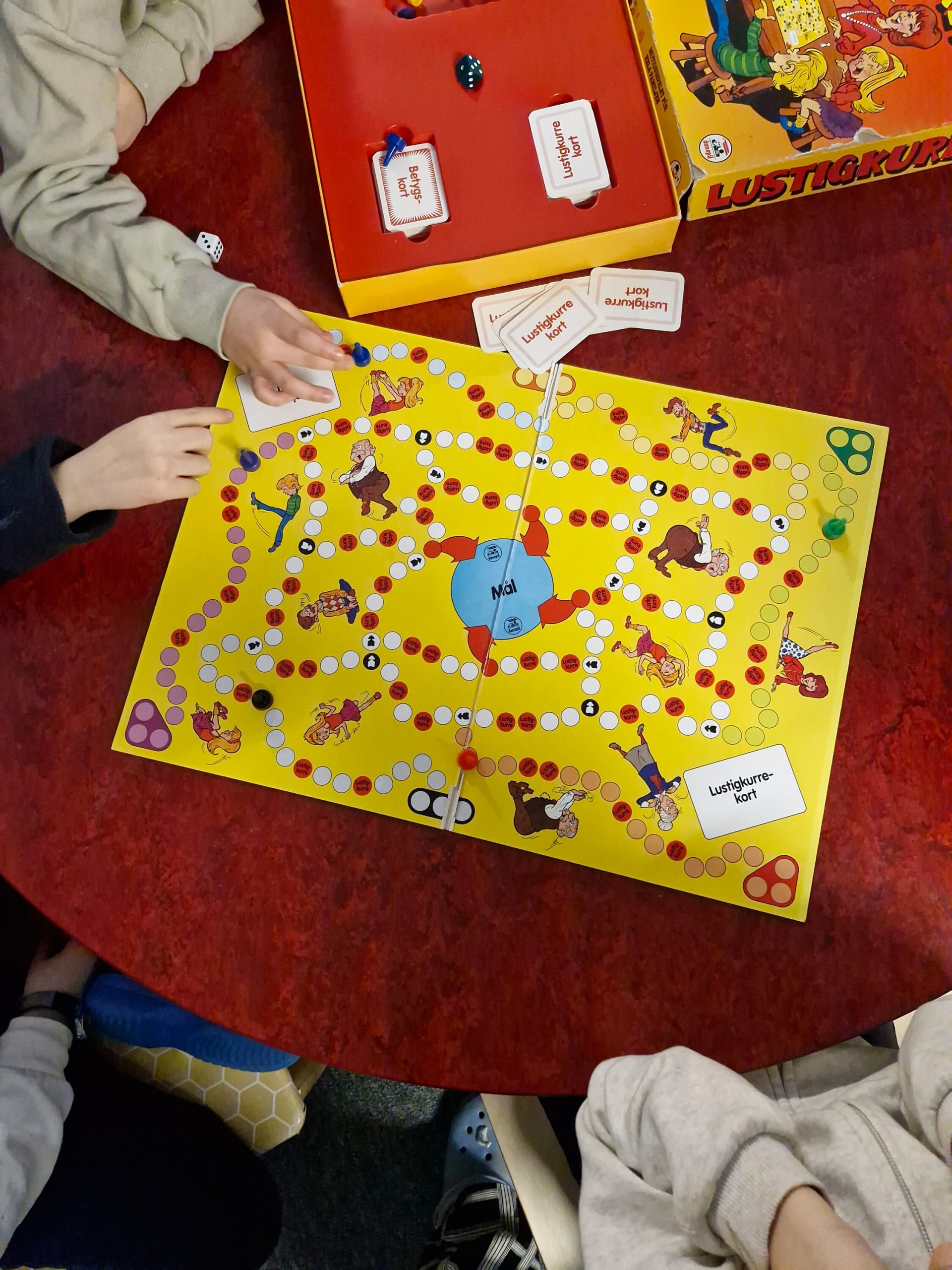 Bild uppifrån av barn som spelar brädspelet Lustigkurre vid ett runt bord, med spelpjäser, tärning och kort synliga på spelbrädet.