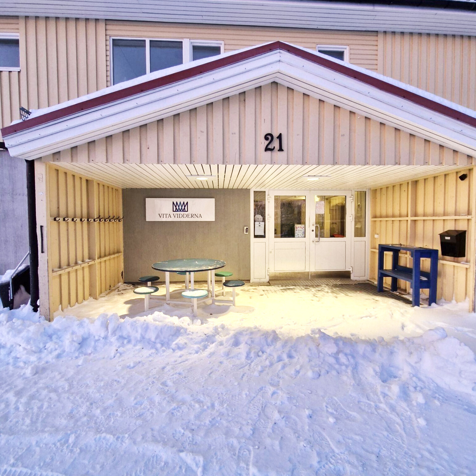 Snötäckt entré till en skolbyggnad med beige panel och rött takfog. Under ett skärmtak med nummer 21 syns en skylt med texten 'Vita Vidderna' och en kronlogotyp. I entréområdet finns knopar för ytterkläder, ett runt bord med pallar i grönt och vitt, samt vita dubbeldörrar in till byggnaden. Till höger står en blå postlåda på ett bord. Marken framför är täckt av snö.