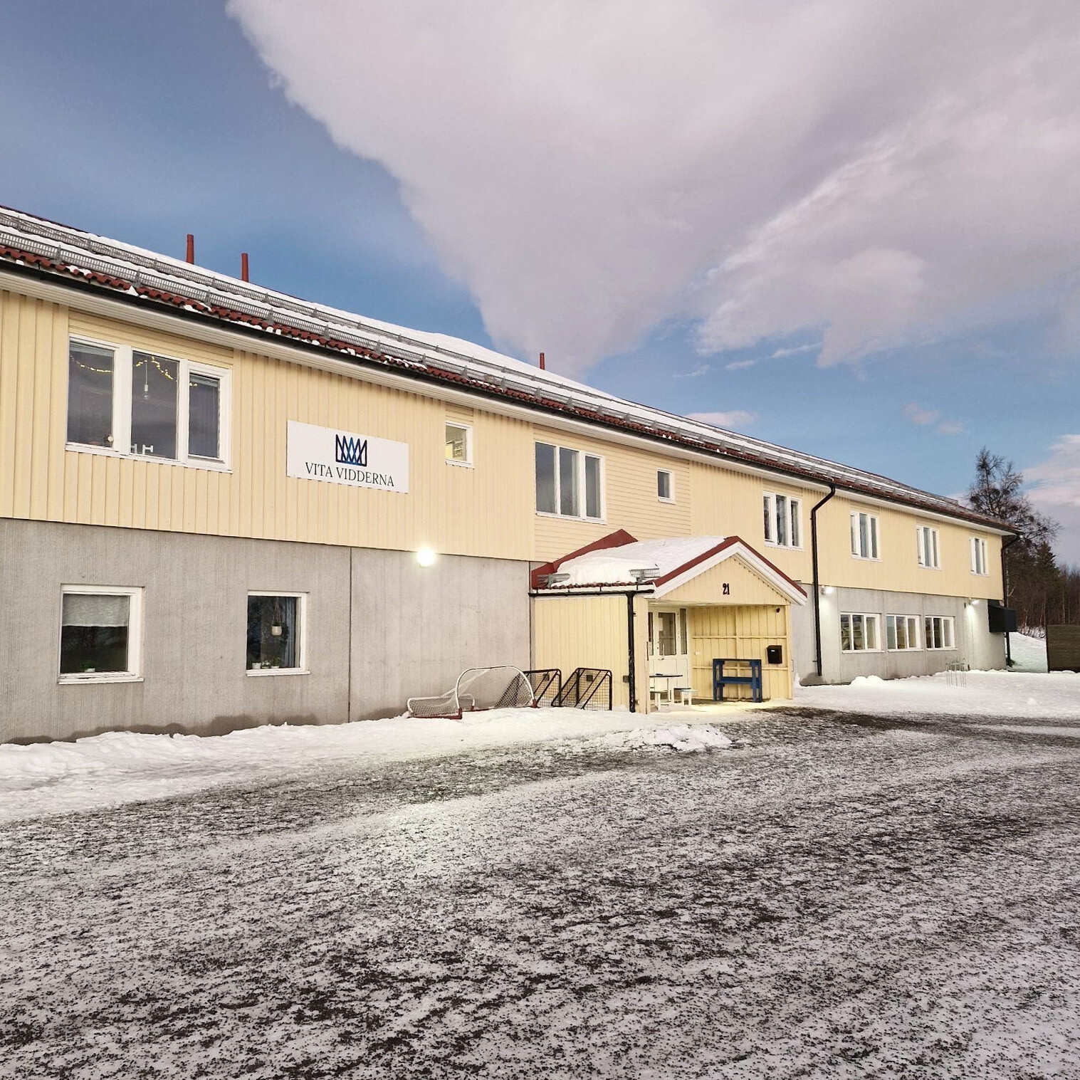 Skolbyggnaden Vita Vidderna med gul fasad, fotograferad i ett vinterlandskap med snö på marken och en dramatisk himmel med moln.
