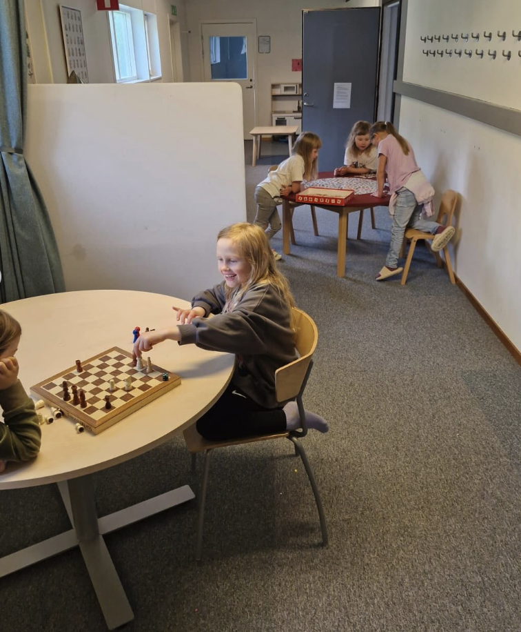 En leende elev spelar schack vid ett runt bord, medan andra elever i bakgrunden spelar ett annat spel vid ett rött bord i en skolkorridor.