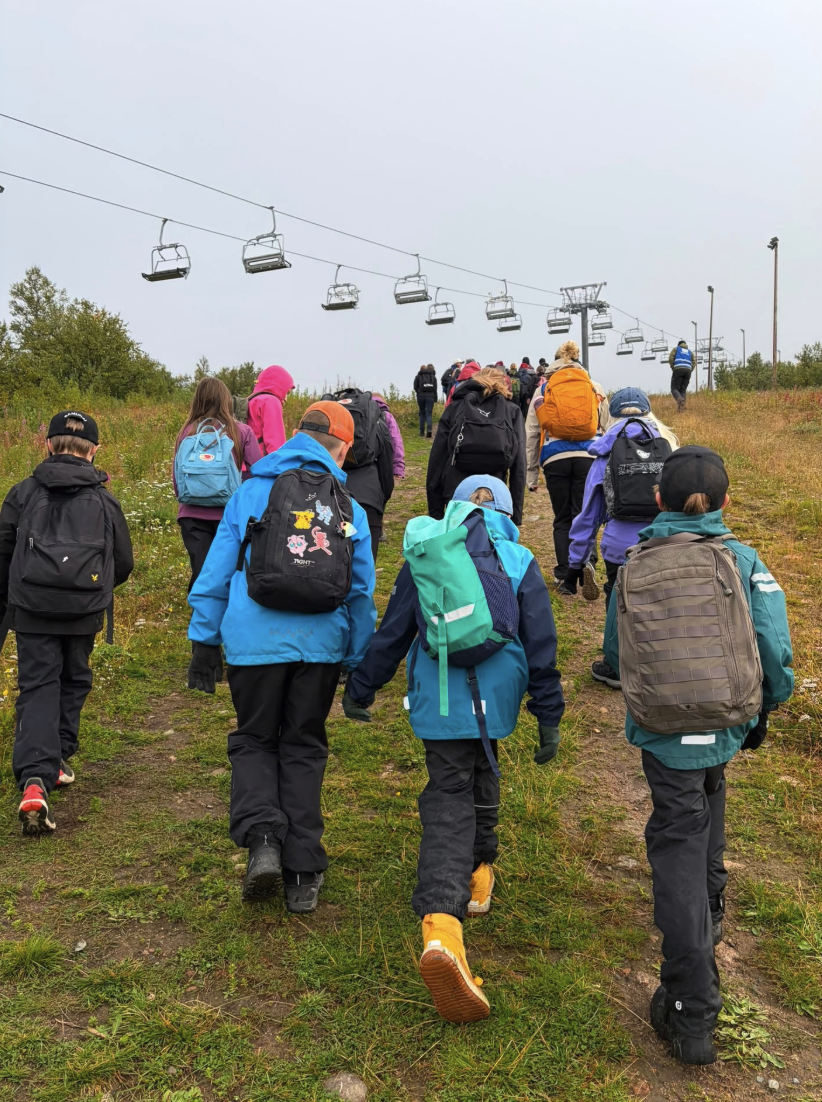 En grupp elever med ryggsäckar vandrar uppför en gräsbevuxen backe under en stillastående stolslift, troligen på ett skidområde utanför säsong.